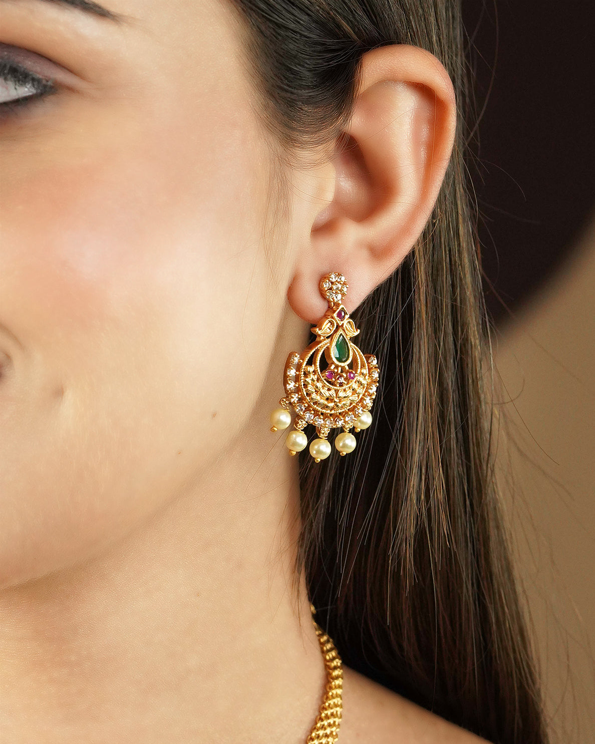 Close-up of a woman's ear wearing a gold earring with intricate design, featuring red and green gemstones and dangling pearls.