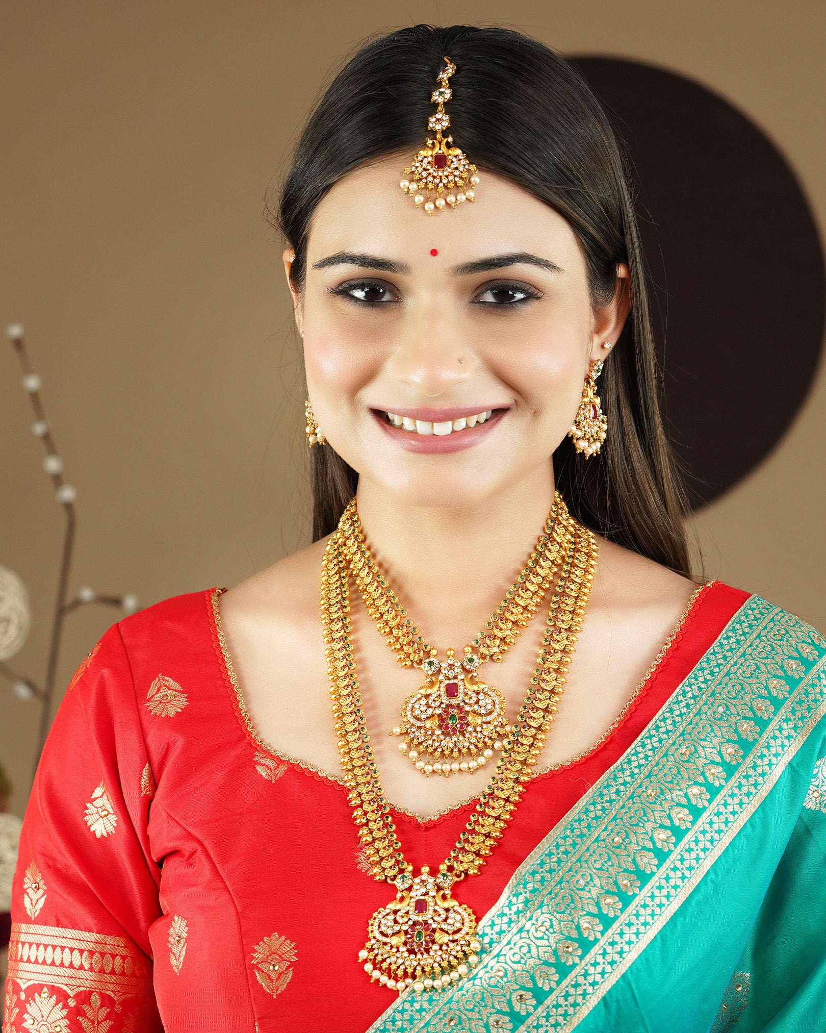 A woman wearing traditional gold jewelry, including a maang tikka, earrings, and layered necklaces, paired with a red and green saree.
