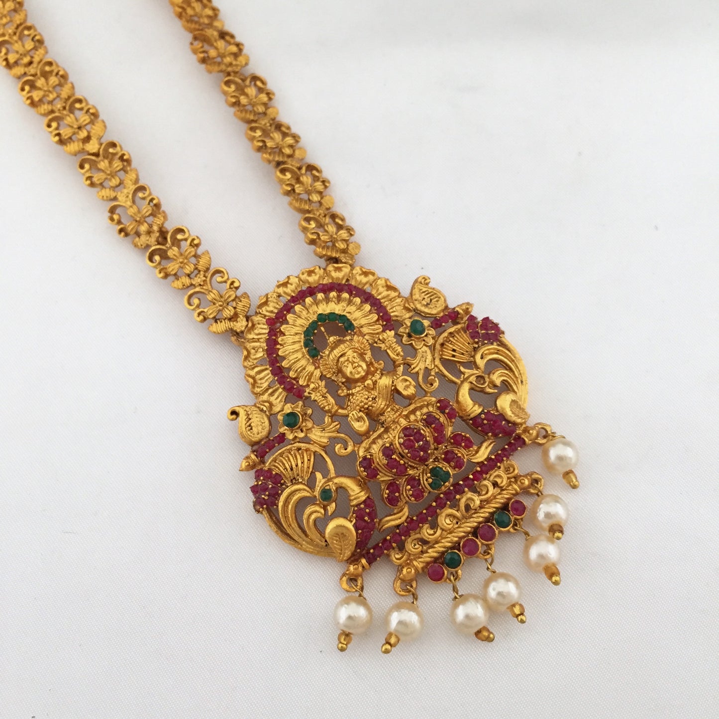 Gold necklace with an intricate pendant featuring a deity, adorned with red and green gemstones and pearl drops.