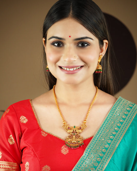 A woman wearing a red and green traditional outfit, adorned with a gold necklace and matching earrings.