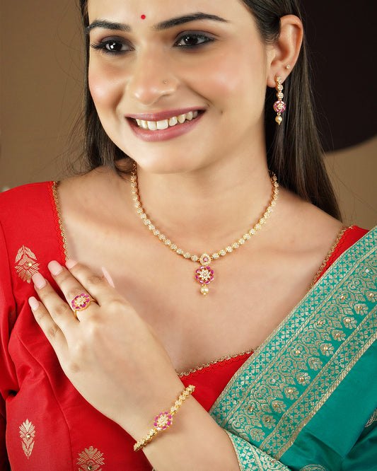 A woman wearing a gold necklace with a pink and white floral pendant, matching earrings, a ring, and a bracelet. She is dressed in a red and green traditional outfit.