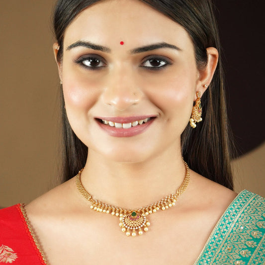 A woman wearing a gold necklace with a central pendant adorned with red and green stones and pearl accents, along with matching gold earrings.