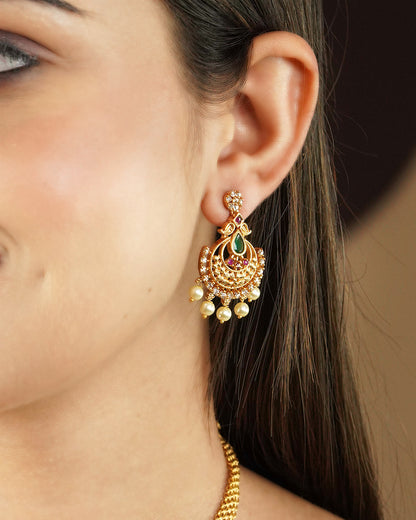 Close-up of a woman's ear wearing a gold earring with intricate design, featuring red and green gemstones and dangling pearls.