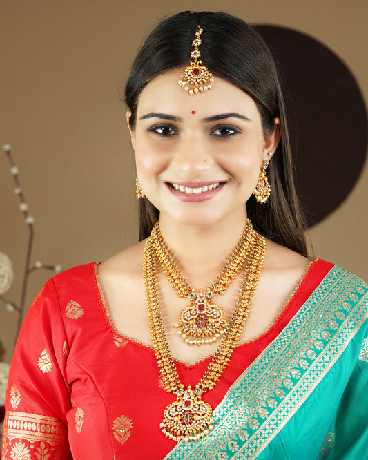 A woman wearing traditional gold jewelry, including a maang tikka, earrings, and layered necklaces, paired with a red and green saree.