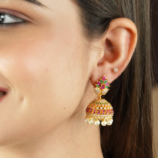 A close-up of a woman's ear wearing a colorful jhumka earring adorned with gemstones and pearl drops.