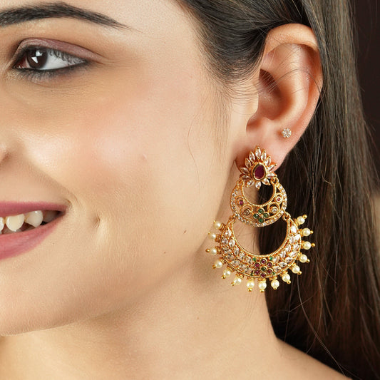 A woman wearing a large, ornate gold earring with red and green gemstones and pearl accents.