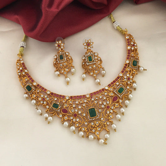 A set of traditional gold jewelry consisting of a necklace and matching earrings. The necklace features intricate designs with green and red gemstones, white pearls, and small white crystals. The earrings have a similar design with green and red gemstones, white pearls, and small white crystals. The jewelry is displayed on a white surface with a maroon fabric in the background.