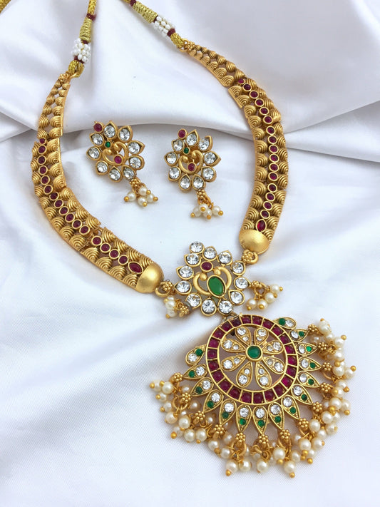 A gold necklace set with intricate designs, featuring a large circular pendant adorned with green, red, and white stones, and matching earrings.