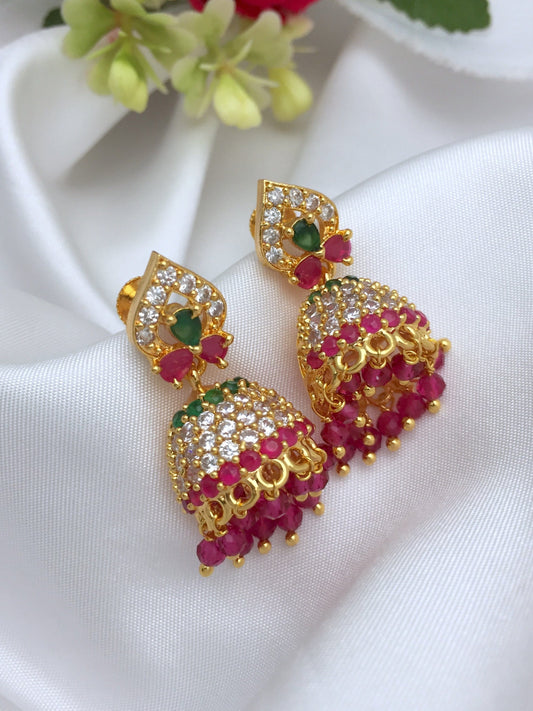 A pair of gold jhumka earrings adorned with red and green gemstones and intricate white stone detailing.
