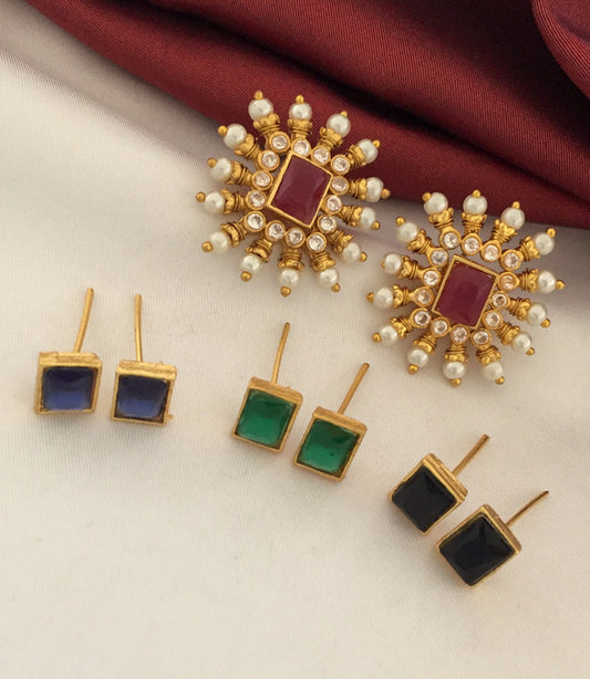 A set of five pairs of earrings displayed on a white surface with a red fabric in the background. The top pair features large, ornate gold earrings with a central red stone, surrounded by smaller white stones and pearls. Below are four pairs of smaller, rectangular stud earrings in blue, green, black, and red.