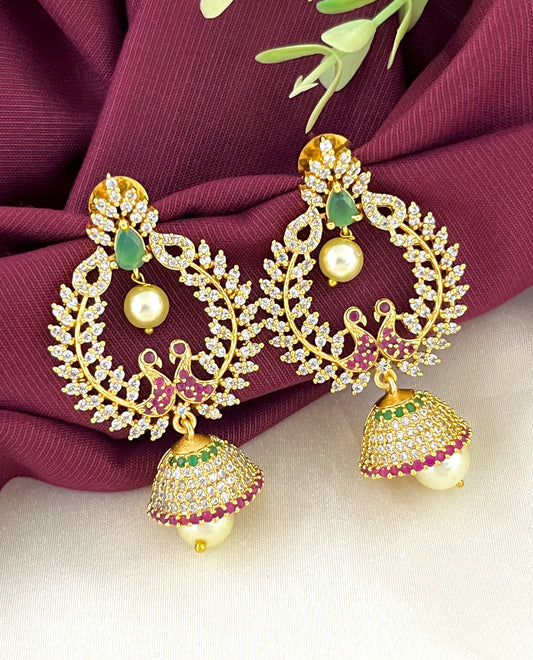 A pair of ornate gold earrings with intricate designs, featuring green and red gemstones, white pearls, and a jhumka-style hanging element.
