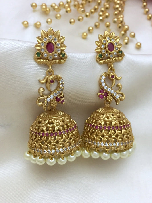 Gold jhumka earrings with intricate peacock design, adorned with CZ stones, red and green gemstones, and pearl drops.